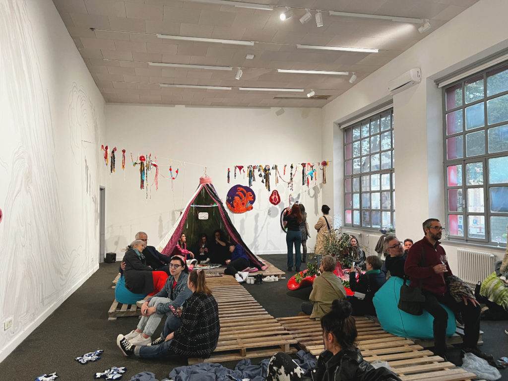 Talaya Schmid Liquid Cuntry Installation 2023. Image shows the opening crowd sitting on fabric cushions in front of a tent.